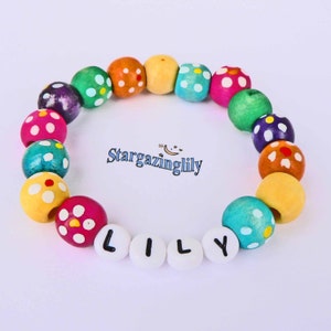 May include: A colorful beaded bracelet with the name "LILY" spelled out in white letter beads. The bracelet features a variety of wooden beads in different colors, including pink, green, yellow, and blue, each with white polka dots. The text "Stargazinglily" is also visible.