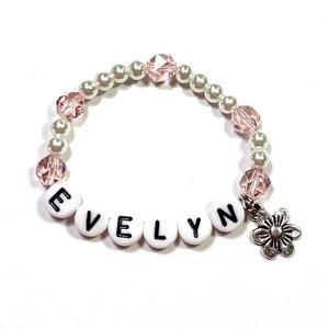 May include: A white and pink beaded bracelet with a silver flower charm and the name "EVELYN" spelled out in black letters on white beads.