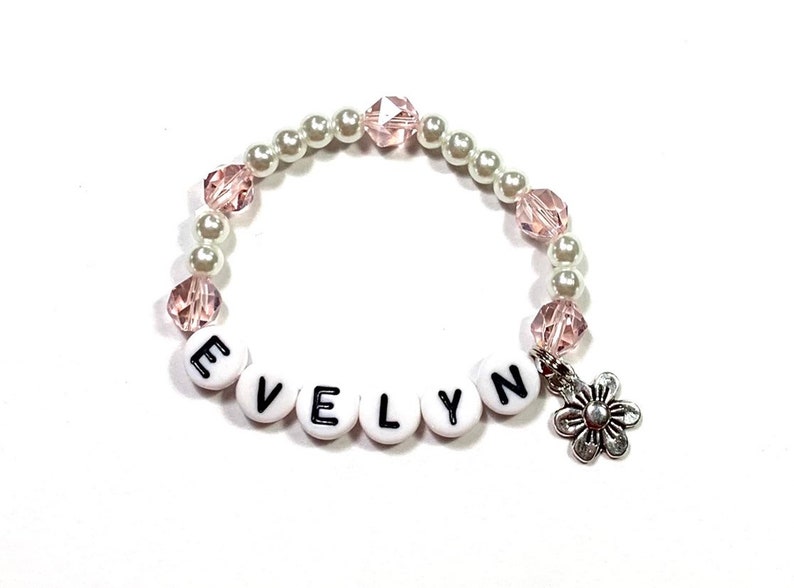 May include: A white and pink beaded bracelet with a silver flower charm and the name "EVELYN" spelled out in black letters on white beads.