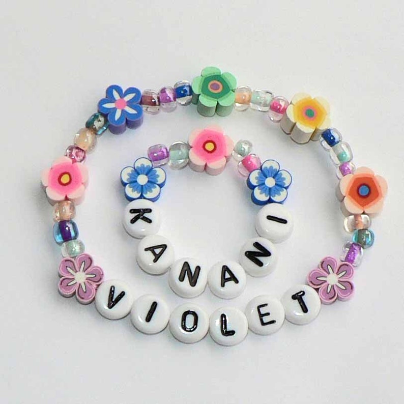 May include: A colorful beaded bracelet with the names "Violet" and "Kanani" spelled out in white beads. The bracelet is decorated with pink, blue, and green flower-shaped beads.