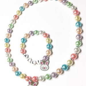 May include: A pastel colored pearl necklace and bracelet set with bunny charms. The necklace has a larger bunny charm and the bracelet has a smaller bunny charm with the word "ALLI" spelled out in white beads.