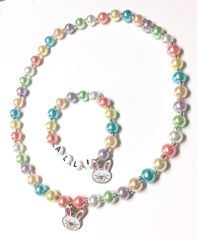 May include: A pastel colored pearl necklace and bracelet set with bunny charms. The necklace has a larger bunny charm and the bracelet has a smaller bunny charm with the word "ALLI" spelled out in white beads.