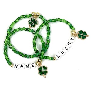May include: Three green beaded bracelets with gold charms. The charms are four-leaf clovers. One bracelet has the word "LUCKY" spelled out in white letters. Another bracelet has the word "NAME" spelled out in white letters.