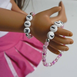 May include: Two beaded bracelets with white beads spelling out the names "MIA" and "LILY" in black letters. The bracelets are on a doll's wrist.
