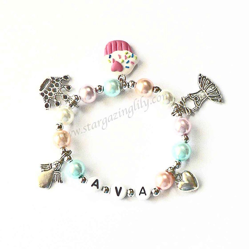 May include: A pink, blue, and white beaded bracelet with silver charms, including a crown, a cupcake, a heart, and an angel. The bracelet also has white letter beads spelling out the name "AVA".