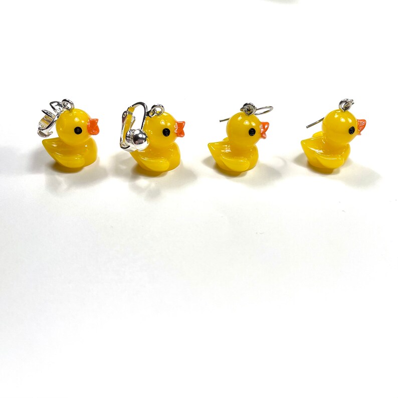 May include: Four yellow rubber duck earrings with orange beaks and black eyes. Two of the earrings are clip-on style and two are dangle earrings.