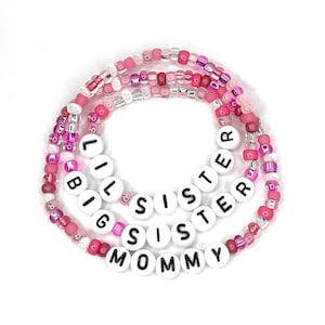 May include: Three pink and white beaded bracelets with the words "Big Sister", "Lil Sister", and "Mommy" spelled out in black letters on white beads.