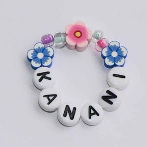 May include: A white beaded bracelet with the name "KANANI" spelled out in black letters. The bracelet is decorated with blue and white flower beads and a pink flower bead with a yellow center.