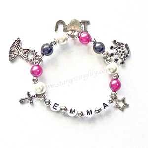 May include: A silver charm bracelet with pink, white, and black beads. The charms include a cross, a star, a crown, a dress, and the letters "EMMA".