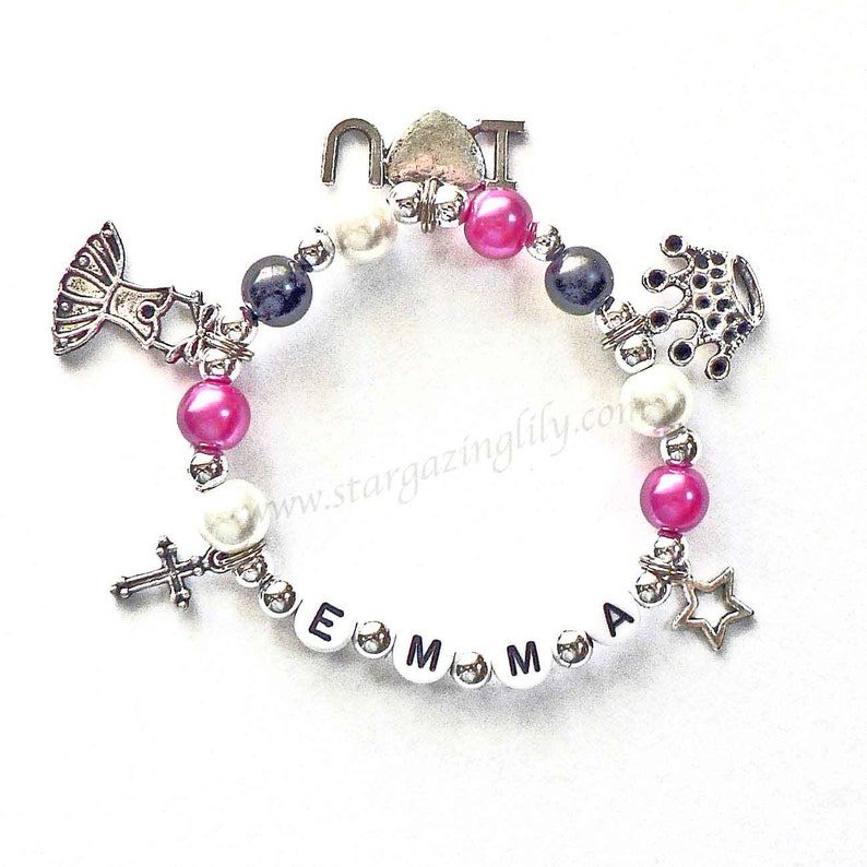 May include: A silver charm bracelet with pink, white, and black beads. The charms include a cross, a star, a crown, a dress, and the letters "EMMA".
