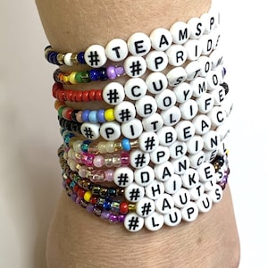 May include: A stack of colorful beaded bracelets with white beads that spell out hashtags and words like #TEAMSP, #PRIDE, #CUSTOM, #BOYMO, #PI, #TLIFE, #BEAC, #PRIN, #DANCEL, #HIKE, #AUT'S, and #LUPUS.