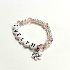 May include: White beaded bracelet with the name "EVELYN" spelled out in black letters. The bracelet has pink and white beads and a silver flower charm.