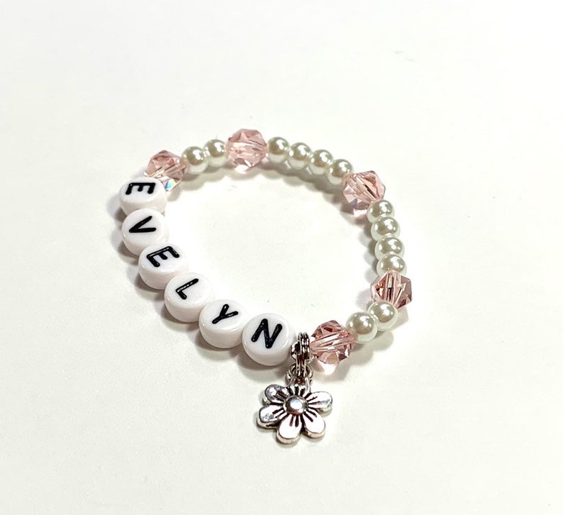 May include: White beaded bracelet with the name "EVELYN" spelled out in black letters. The bracelet has pink and white beads and a silver flower charm.