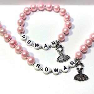 May include: Two pink pearl bracelets with the name "ROWAN" spelled out in white beads and a silver ballerina charm.
