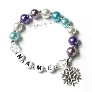 May include: A white beaded bracelet with a snowflake charm and the word "NAME" spelled out in black letters. The beads are purple, white, and blue.