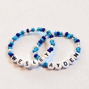 May include: Two beaded bracelets with the names "Wesley" and "Ayden" spelled out in white beads with black lettering. The bracelets are made with blue and white beads.