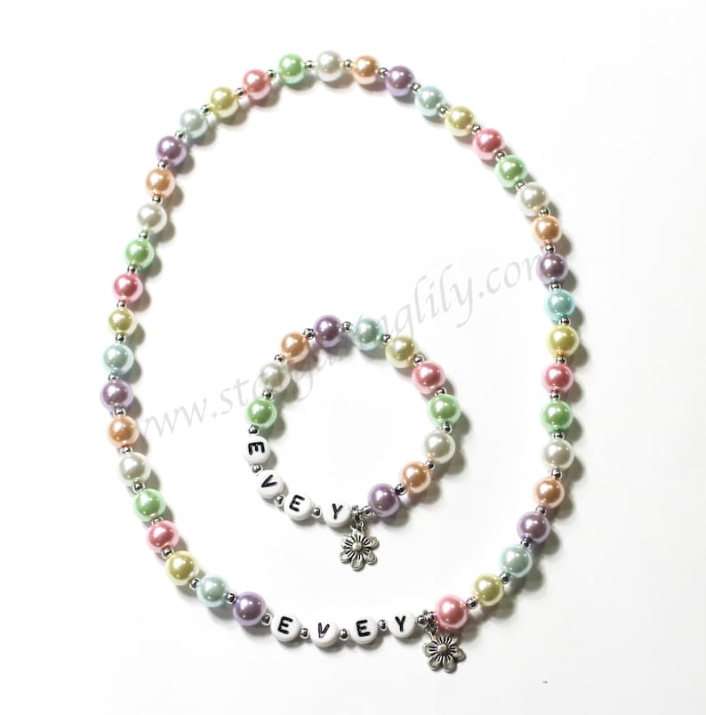 May include: A pastel-colored pearl necklace and bracelet set with a flower charm and the name "EVEY" spelled out in white letters.