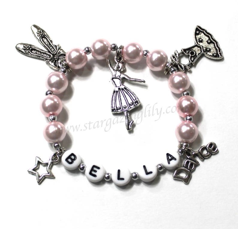 May include: A pink beaded bracelet with silver charms, including a ballerina, a star, a shoe, a dress, and the word "Dance". The bracelet also has white beads with the letters "BELLA" on them.