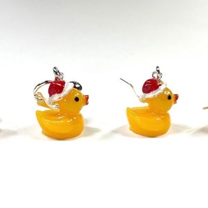 May include: Four pairs of yellow rubber ducky earrings with red and white Santa hats. The earrings are silver and have a hook closure.