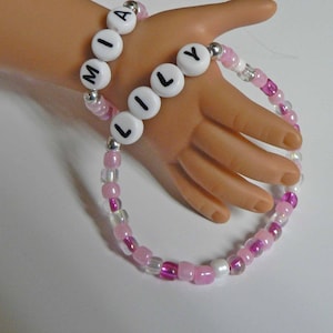 May include: Two beaded bracelets with the names "MIA" and "LILY" spelled out in white letters on white beads. The bracelets are made with pink and clear beads.