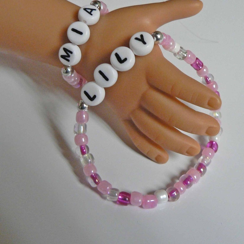 May include: Two beaded bracelets with the names "MIA" and "LILY" spelled out in white letters on white beads. The bracelets are made with pink and clear beads.