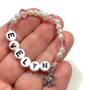 May include: A white and pink beaded bracelet with the name "EVELYN" spelled out in white beads. The bracelet has a silver flower charm.