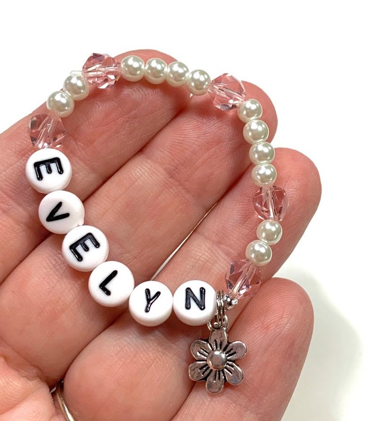May include: A white and pink beaded bracelet with the name "EVELYN" spelled out in white beads. The bracelet has a silver flower charm.