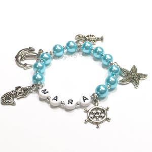 May include: A light blue beaded bracelet with silver charms, including a mermaid, anchor, starfish, and a ship's wheel. The bracelet also has white beads with the name "MARA" spelled out.