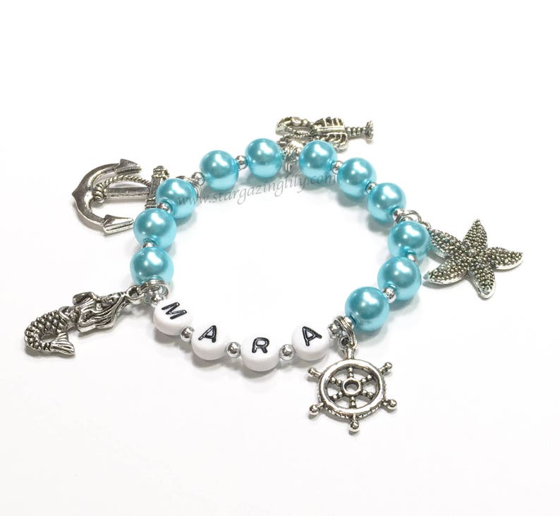 May include: A light blue beaded bracelet with silver charms, including a mermaid, anchor, starfish, and a ship's wheel. The bracelet also has white beads with the name "MARA" spelled out.