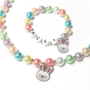 May include: A pastel rainbow colored beaded necklace and bracelet set with bunny charms. The bracelet has the name "ALLI" spelled out in white letters.