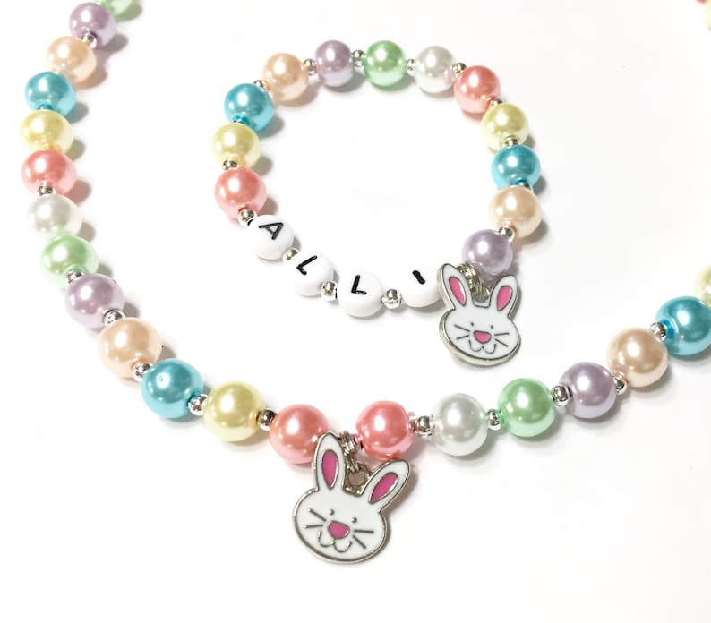 May include: A pastel rainbow colored beaded necklace and bracelet set with bunny charms. The bracelet has the name "ALLI" spelled out in white letters.