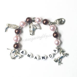 May include: A pink and brown beaded bracelet with silver charms, including a horse head, a cowboy hat, a boot, and two horses. The bracelet has white beads with the name "Amanda" spelled out.