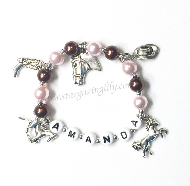 May include: A pink and brown beaded bracelet with silver charms, including a horse head, a cowboy hat, a boot, and two horses. The bracelet has white beads with the name "Amanda" spelled out.
