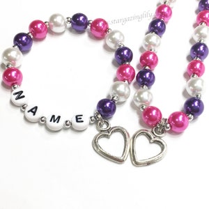 May include: Two beaded necklaces with pink, white and purple beads. One necklace has the word "NAME" spelled out with white letter beads. Both necklaces have silver heart charms.