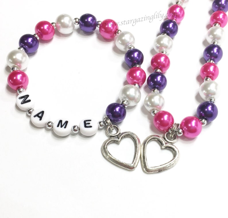May include: Two beaded necklaces with pink, white and purple beads. One necklace has the word "NAME" spelled out with white letter beads. Both necklaces have silver heart charms.