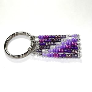 May include: A silver key ring with a purple, lavender, and white beaded tassel. The beads are arranged in a gradient pattern.