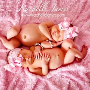 May include: Two newborn babies sleeping on a pink fluffy blanket. Both babies are wearing pink headbands with bows. The babies are lying next to each other, with one baby's arm around the other baby's waist.