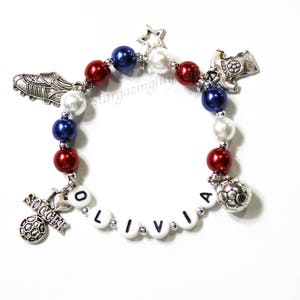 May include: A red, white, and blue beaded bracelet with silver charms, including a soccer ball, a soccer jersey, a soccer cleat, a star, and the name "Olivia".