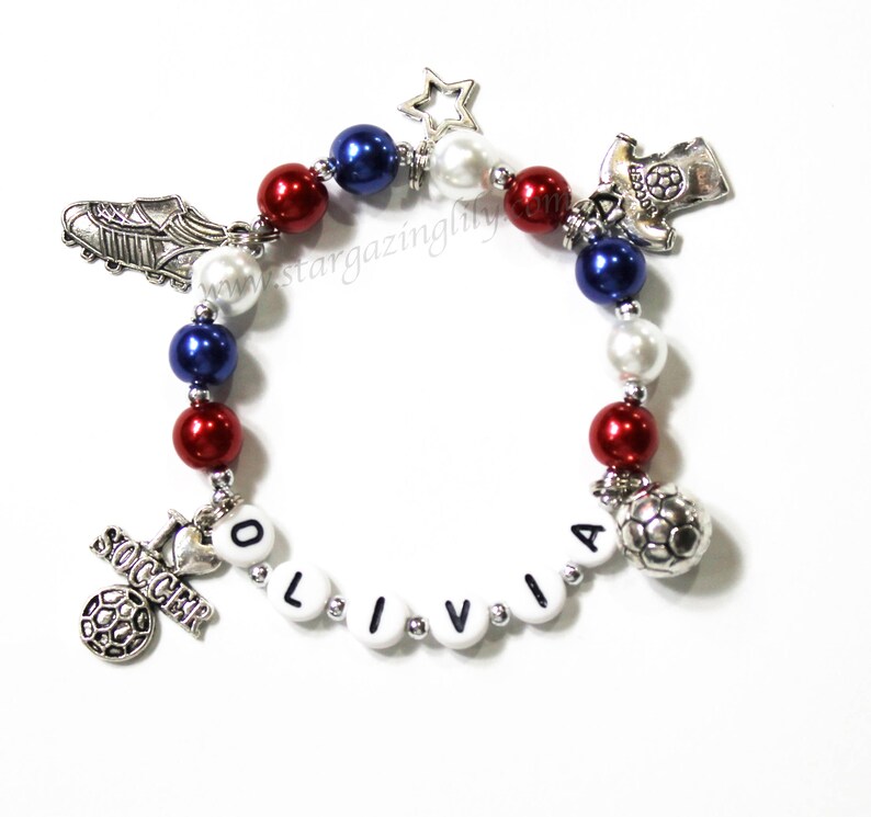May include: A red, white, and blue beaded bracelet with silver charms, including a soccer ball, a soccer jersey, a soccer cleat, a star, and the name "Olivia".