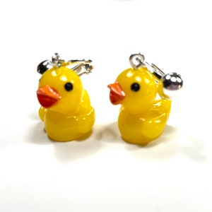 May include: A pair of yellow rubber ducky earrings with silver clip-on backs. The ducks have black eyes and orange beaks.