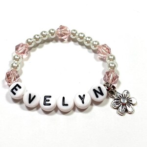 May include: A white and pink beaded bracelet with the name "EVELYN" spelled out in black letters on white beads. A silver flower charm hangs from the bracelet.
