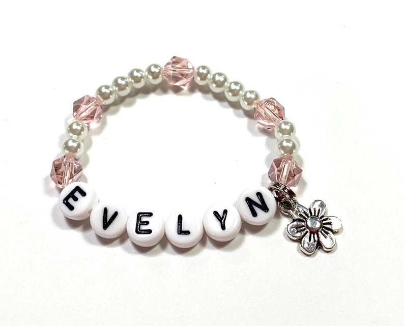 May include: A white and pink beaded bracelet with the name "EVELYN" spelled out in black letters on white beads. A silver flower charm hangs from the bracelet.