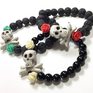 May include: Three black lava rock bracelets with skull and crossbones charms and flower accents. The bracelets are layered on a white surface.
