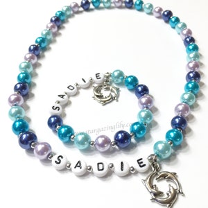 May include: A blue and purple beaded necklace with a silver dolphin charm and the name "SADIE" spelled out in white beads.