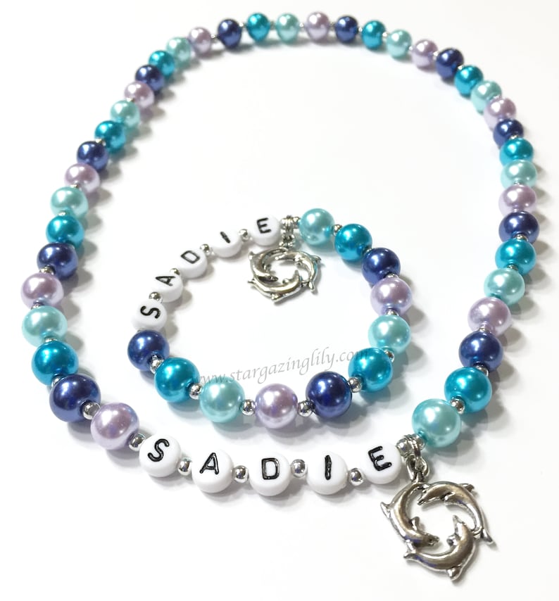 May include: A blue and purple beaded necklace with a silver dolphin charm and the name "SADIE" spelled out in white beads.