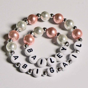 May include: A white and pink beaded bracelet with the name "BAILEY" spelled out in black letters on white beads.