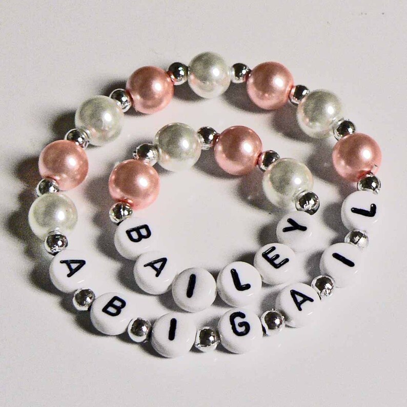 May include: A white and pink beaded bracelet with the name "BAILEY" spelled out in black letters on white beads.