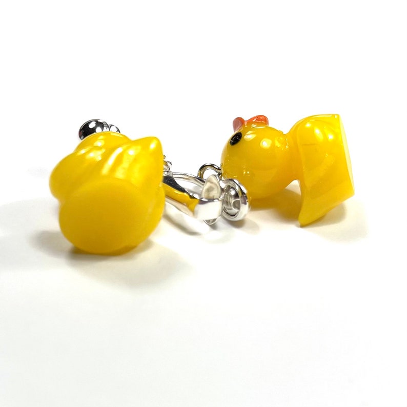 May include: A pair of yellow rubber ducky clip-on earrings. The earrings are made of a yellow rubber material and feature a small, detailed duck design. The earrings are attached to a silver clip-on earring.