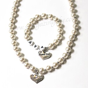 May include: A white pearl necklace and bracelet set with a silver heart charm that says "Flower Girl". The bracelet has white letter beads that spell "AVA".