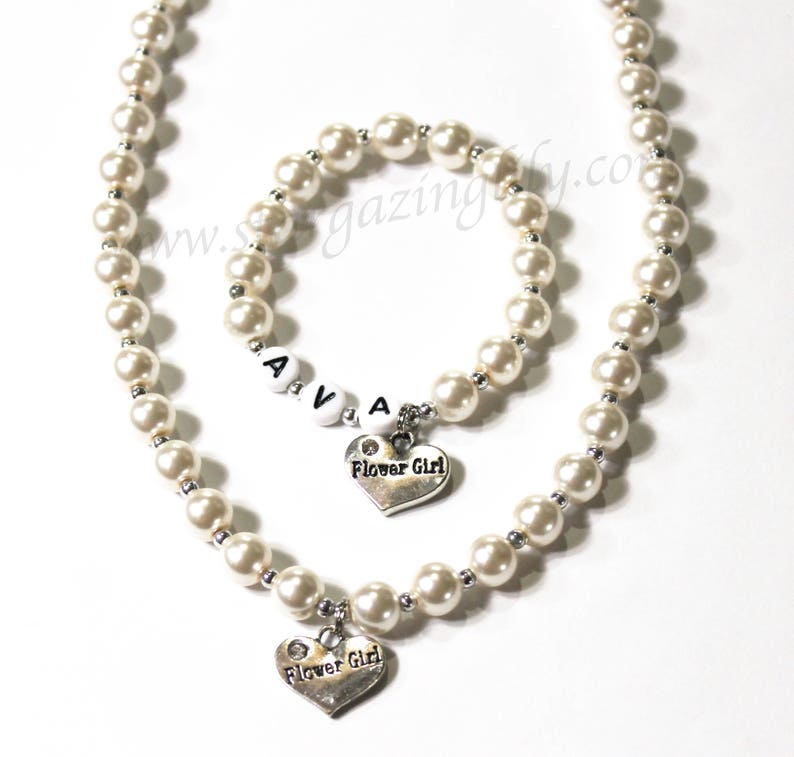 May include: A white pearl necklace and bracelet set with a silver heart charm that says "Flower Girl". The bracelet has white letter beads that spell "AVA".